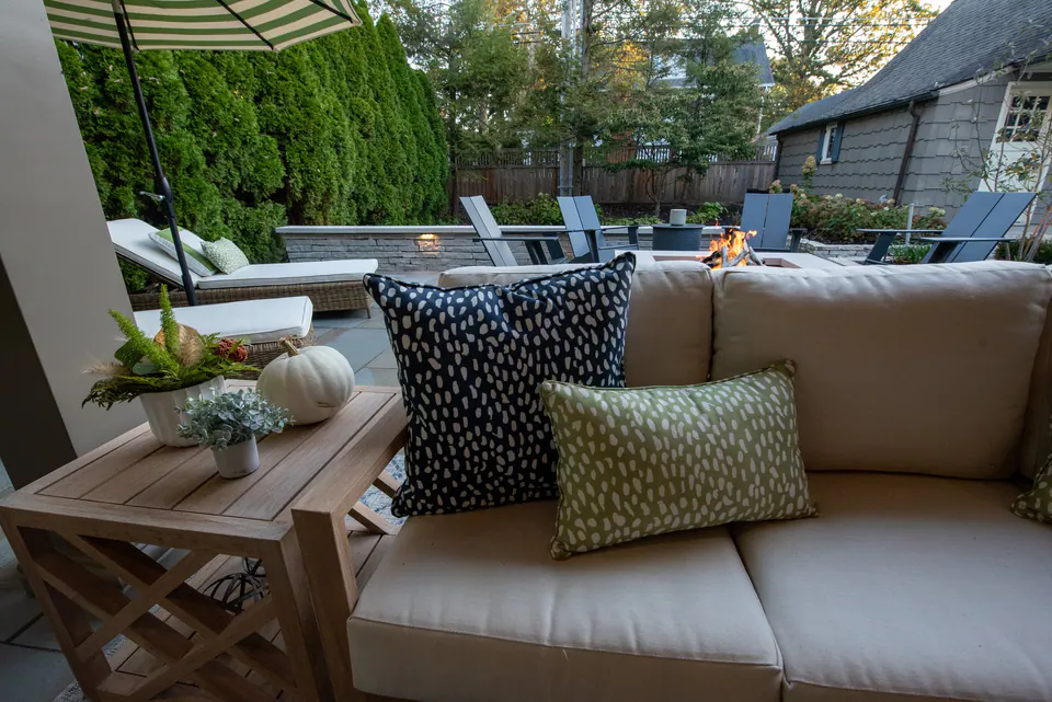 Covered lounge sofa with pillows looking toward square fire feature, umbrella, paver terrace, and privacy planting